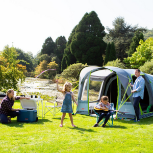 an image showing a family platying outside a tent on green grass with a background of a lake and adult trees. The family are positioned infront of a family sized tent . 