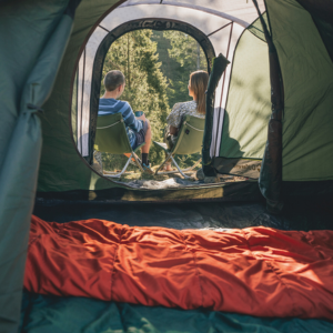 An image showing a tent shot from the inside and a couple sitting outside the tent opening on green camping chairs looking at a forest view