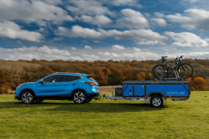 An image showing a green grass and a cloudy blue skie with a 4x4 blue car towing the opus trailer tent folded together with a bike on the back