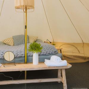 an image showing a traditional canvas tipi tent stu;ed with a rustic bench, mirrow and vase of stems. In the background there is a large double bed, with yellow and blue spotted cushions and  lampshade hung in the middle. 