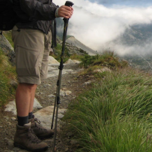 An image showing a man on a hiking trail wearing shorts, hiking boots and holding a hiking pole looking at the view below. 