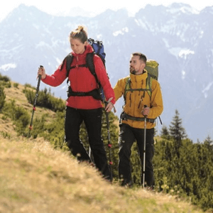 A couple on a heavily inclined trail with mountains in the background hiking in a waterproof jacket, rucksack and hiking poles. 