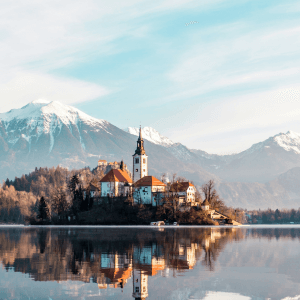 a scene depicting lake bled with snow topped mountains in the background and sun shing on the trees in a winters afternoon